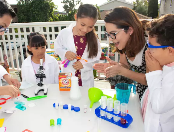 Two adults and three children in lab coats engage in a science experiment outdoors, using various lab equipment and materials.