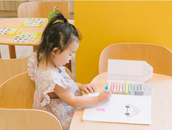 A young child drawing with markers at a table in a brightly colored classroom.