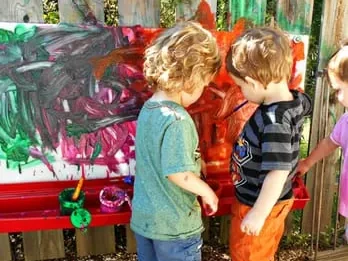 Two young children engaging in painting on a large paper mounted on an outdoor fence, surrounded by paint supplies.